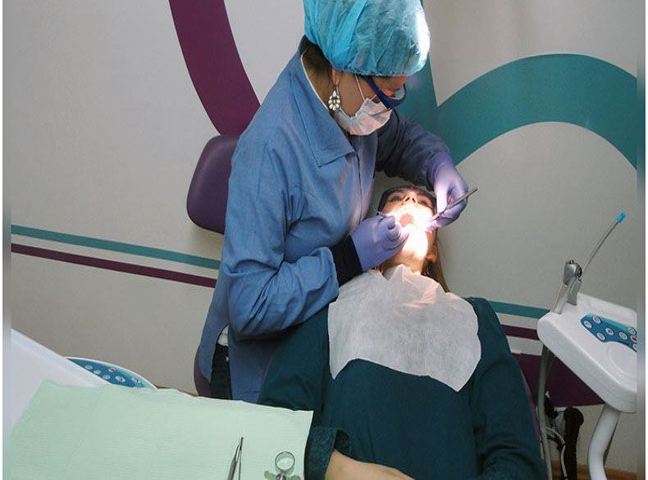 Dentista infantil en España - Un dentista con guantes, mascarilla y cubrepelo examina la boca de un paciente en una clínica dental. El paciente está tumbado en el sillón dental con un babero, y en primer plano se ven instrumentos dentales. | Clínica dental pediátrica Dentistainfantilcercademi