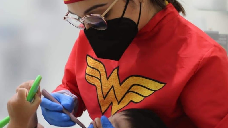 Dentista infantil en España - Una dentista vestida con una bata roja con el logotipo dorado de Wonder Woman y una máscara negra atiende a un paciente, utilizando instrumentos dentales con las manos enguantadas. El paciente sostiene un espejo verde para observar. | Clínica dental pediátrica Dentistainfantilcercademi