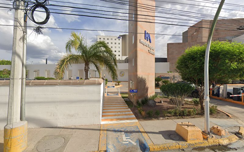 Dentista infantil en España - Vista de la entrada de un hospital con un letrero alto, una palmera, un paso de peatones y una rampa accesible para sillas de ruedas, ideal para visitas a una odontopediatra en Torreón. El edificio beige presenta un logotipo azul, con árboles y líneas eléctricas en primer plano. | Clínica dental pediátrica Dentistainfantilcercademi