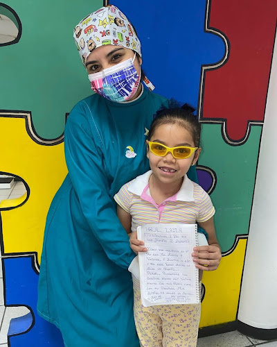 Dentista infantil en España - Una mujer con bata médica y una colorida mascarilla abraza a una sonriente niña con gafas amarillas, que sostiene una nota manuscrita. Están delante de un colorido muro con piezas de puzzle. | Clínica dental pediátrica Dentistainfantilcercademi