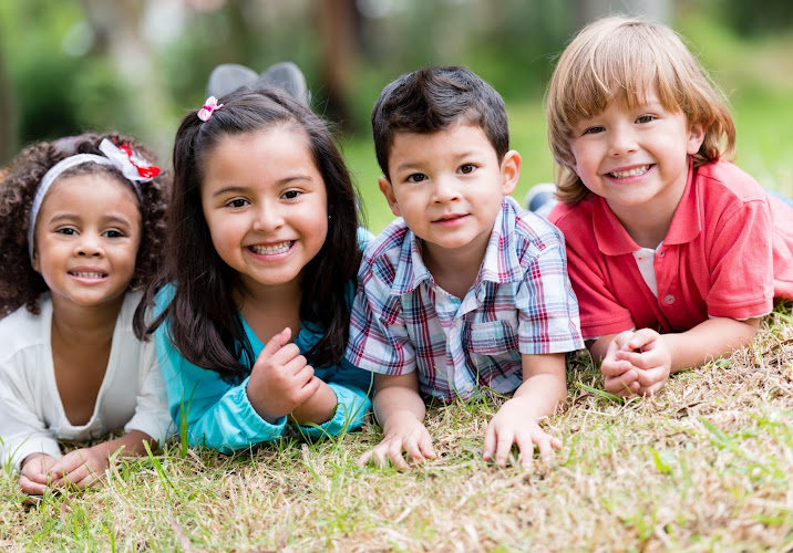 Dentista infantil en España - Cuatro niños pequeños tumbados sobre la hierba sonríen a la cámara. Están muy juntos, disfrutando de un día soleado al aire libre. El fondo está difuminado con árboles verdes, creando un ambiente alegre y juguetón. | Clínica dental pediátrica Dentistainfantilcercademi