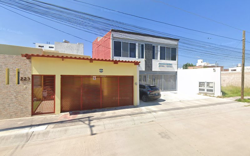 Dentista infantil en España - Vista de una calle de una zona residencial en la que se ve una casa amarilla con una verja roja, un coche negro aparcado junto a un edificio blanco y gris de dos plantas y cables de alta tensión en lo alto en un día soleado. | Clínica dental pediátrica Dentistainfantilcercademi