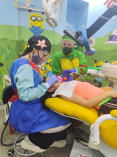 Dentista infantil en España - Una niña está tumbada en un sillón dental mientras dos dentistas con batas de colores y protectores faciales la atienden. La habitación tiene paredes brillantes con personajes de dibujos animados y equipos dentales alrededor. | Clínica dental pediátrica Dentistainfantilcercademi