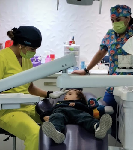 Dentista infantil en España - Un niño se tumba en un sillón dental mientras un dentista con bata amarilla le examina. Otro miembro del personal, con bata de colores y mascarilla, observa en una clínica moderna y luminosa. | Clínica dental pediátrica Dentistainfantilcercademi