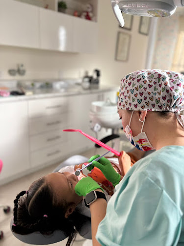 Dentista infantil en España - Un dentista con gorro y mascarilla con motivos de corazones examina los dientes de una niña en una luminosa clínica dental. La niña se recuesta en el sillón dental sosteniendo un espejo rosa. | Clínica dental pediátrica Dentistainfantilcercademi