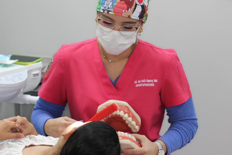 Dentista infantil en España - Un dentista con bata rosa y gorro de colores muestra a un paciente reclinado las técnicas de cepillado con un gran modelo de dientes. | Clínica dental pediátrica Dentistainfantilcercademi