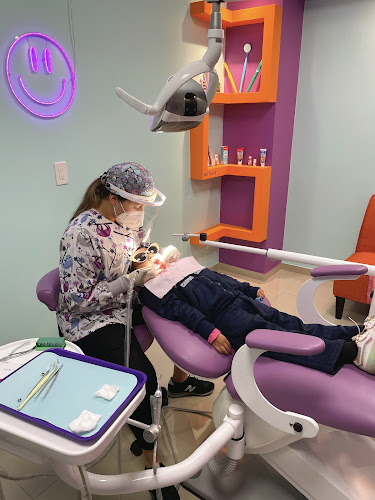 Dentista infantil en España - Un dentista con equipo de protección examina a un niño en una colorida clínica dental con sillones morados, estanterías naranjas y un letrero de neón con una cara sonriente en la pared. Las herramientas dentales están dispuestas en una bandeja cercana. | Clínica dental pediátrica Dentistainfantilcercademi