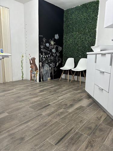 Dentista infantil en España - Una habitación moderna con suelo de imitación madera, dos sillas blancas junto a una pared con vegetación artificial, una pizarra con dibujos infantiles y armarios blancos a la derecha. | Clínica dental pediátrica Dentistainfantilcercademi