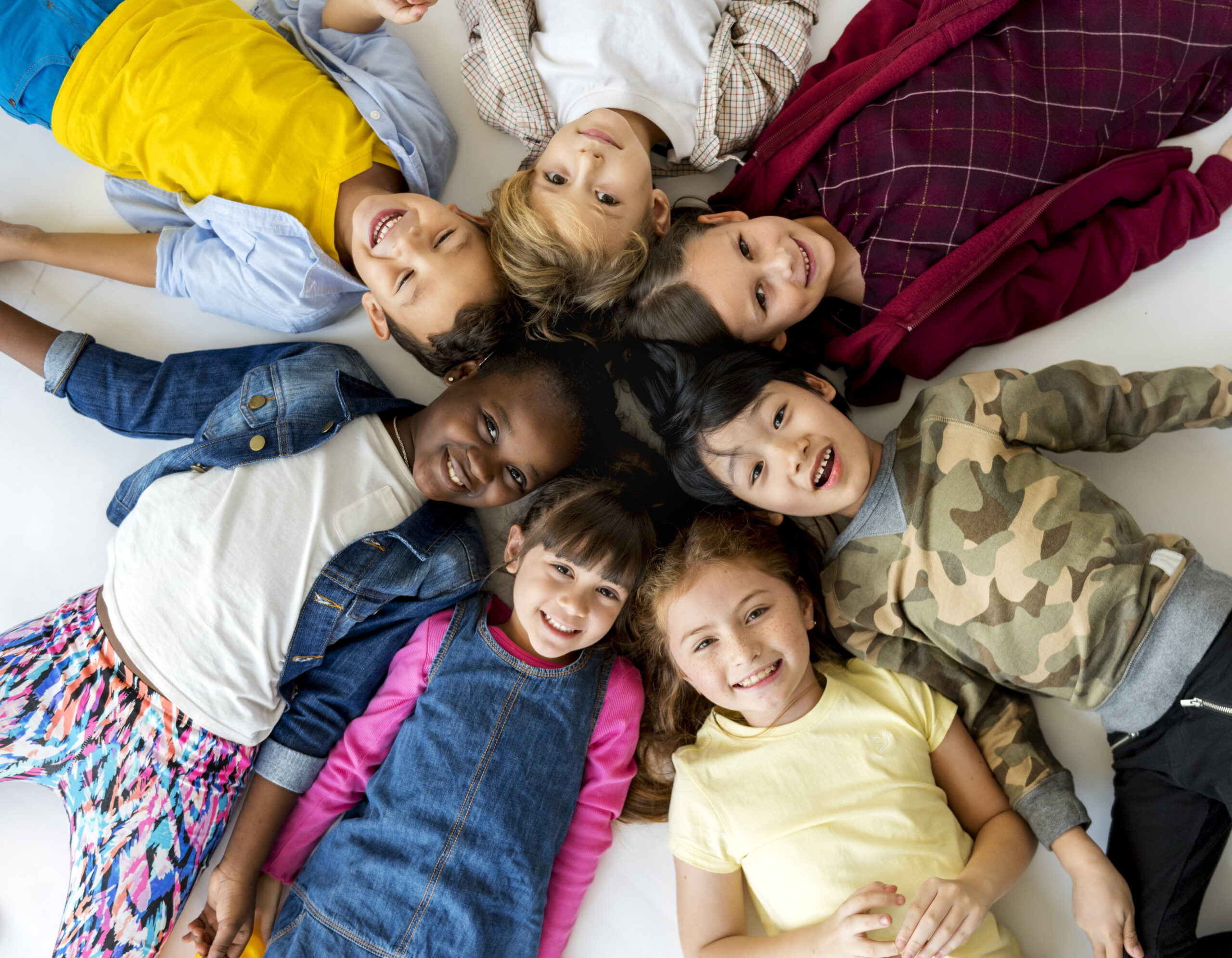 Dentista infantil en España - Un grupo de siete niños diversos están tumbados en el suelo formando un círculo, con las cabezas juntas y sonriendo a la cámara sobre un fondo blanco. Llevan ropa informal de colores y parecen felices y relajados. | Clínica dental pediátrica Dentistainfantilcercademi