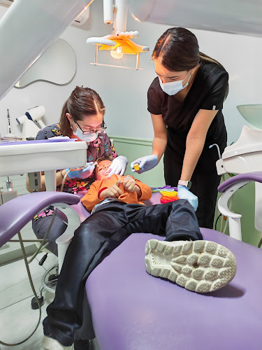 Dentista infantil en España - Un niño se tumba en un sillón dental morado mientras un dentista le examina la boca y un auxiliar le ayuda. Ambos adultos llevan mascarillas y guantes. El niño sostiene un juguete y parece tranquilo bajo la luz dental. | Clínica dental pediátrica Dentistainfantilcercademi
