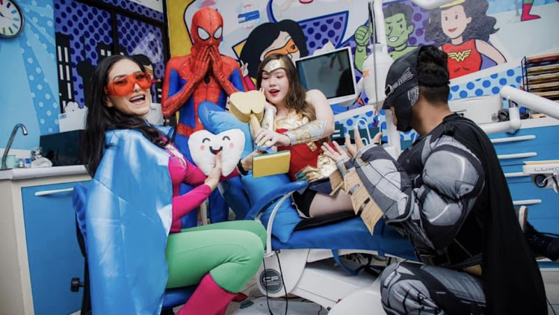 Dentista infantil en España - Cuatro adultos disfrazados de superhéroes, como Spiderman, Wonder Woman y Batman, posan en una colorida consulta dental, sosteniendo un recorte de diente y accesorios dentales, creando un ambiente divertido y animado. | Clínica dental pediátrica Dentistainfantilcercademi