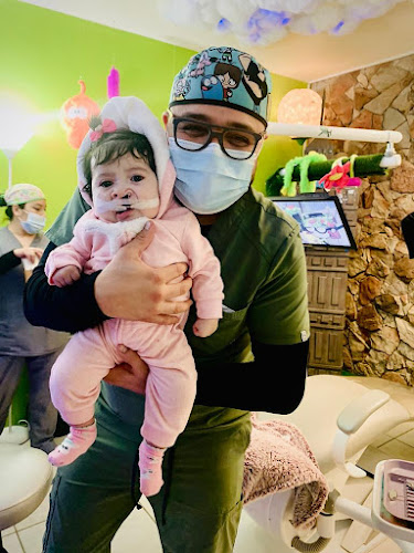Dentista infantil en España - Un dentista sonriente con bata verde y mascarilla sostiene a un bebé vestido de rosa con un vendaje en la cara. La habitación está llena de coloridos adornos lúdicos y otra persona enmascarada está de pie en el fondo. | Clínica dental pediátrica Dentistainfantilcercademi