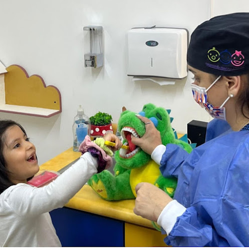 Dentista infantil en España - Una niña practica el cepillado de dientes con un dinosaurio verde de juguete mientras un profesional de la odontología con bata y mascarilla la guía en una colorida consulta dental. | Clínica dental pediátrica Dentistainfantilcercademi