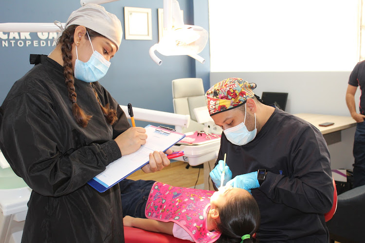 Dentista infantil en España - Un dentista examina los dientes de una niña mientras un ayudante con mascarilla y la cabeza cubierta toma notas en un portapapeles en una clínica dental muy iluminada. | Clínica dental pediátrica Dentistainfantilcercademi
