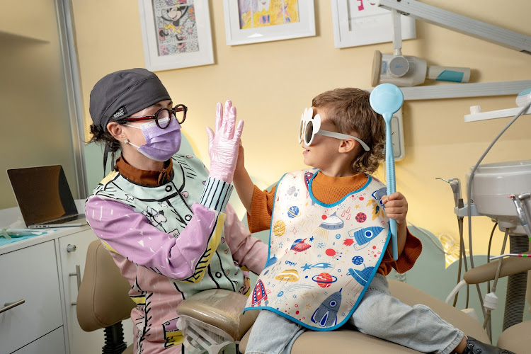 Dentista infantil en España - Una odontopediatra en León con un colorido atuendo y un niño pequeño con gafas de gran tamaño y un divertido babero se chocan los cinco sentados en una alegre consulta dental. | Clínica dental pediátrica Dentistainfantilcercademi