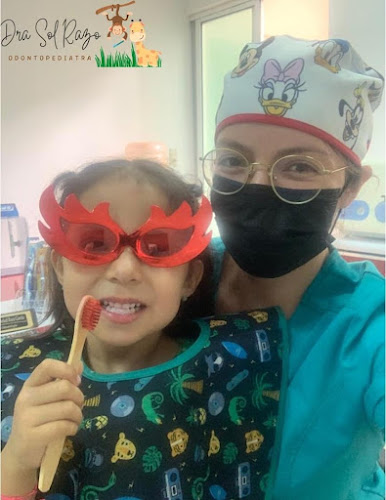 Dentista infantil en España - Un niño sonriente con gafas rojas de novedad y un babero dental sostiene un cepillo de dientes, posando con un odontopediatra en Irapuato con bata, gafas y mascarilla. En el fondo se ve una decoración de animales de dibujos animados. | Clínica dental pediátrica Dentistainfantilcercademi