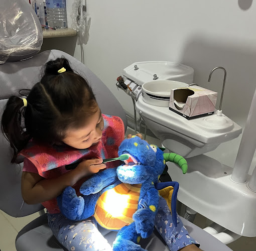 Dentista infantil en España - Una niña sentada en el sillón del dentista sonríe mientras practica el cepillado de los dientes de un dragón de peluche azul. Al fondo se ven instrumentos dentales y un lavabo. | Clínica dental pediátrica Dentistainfantilcercademi