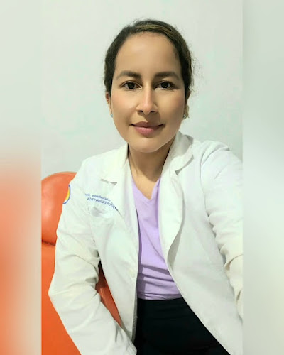 Dentista infantil en España - Una mujer de pelo oscuro, con bata blanca de laboratorio y camisa morada clara, se sienta en una silla naranja y sonríe a la cámara sobre un fondo claro liso. | Clínica dental pediátrica Dentistainfantilcercademi
