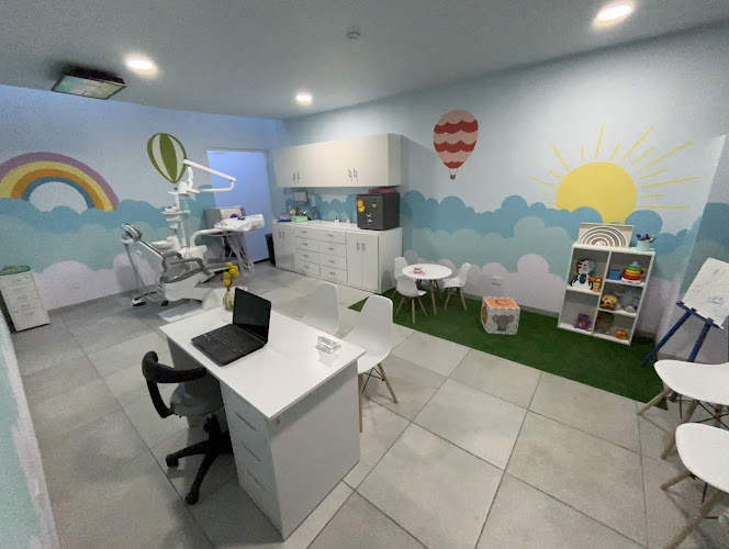 Dentista infantil en España - Un consultorio dental luminoso y acogedor para los niños, con coloridas ilustraciones en la pared de un arco iris, un sol y globos aerostáticos, con equipos dentales, un escritorio con un ordenador portátil y una zona de juegos con juguetes y estanterías. | Clínica dental pediátrica Dentistainfantilcercademi