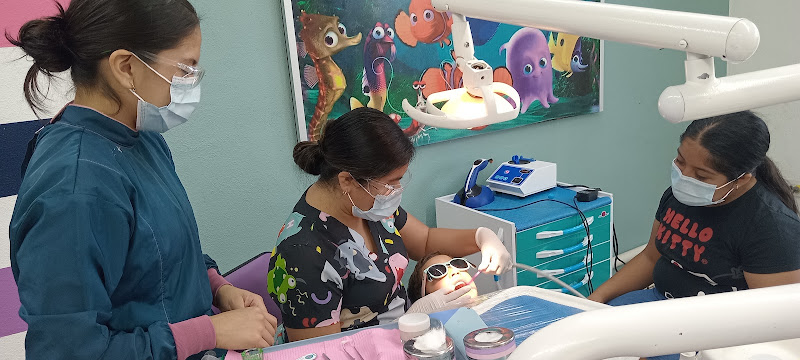 Dentista infantil en España - Un niño con gafas de sol se sienta en un sillón dental mientras un dentista le examina la boca. Dos mujeres, entre ellas una odontopediatra en Irapuato, permanecen de pie cerca. En la pared de la sala hay coloridos adornos de temática acuática. | Clínica dental pediátrica Dentistainfantilcercademi
