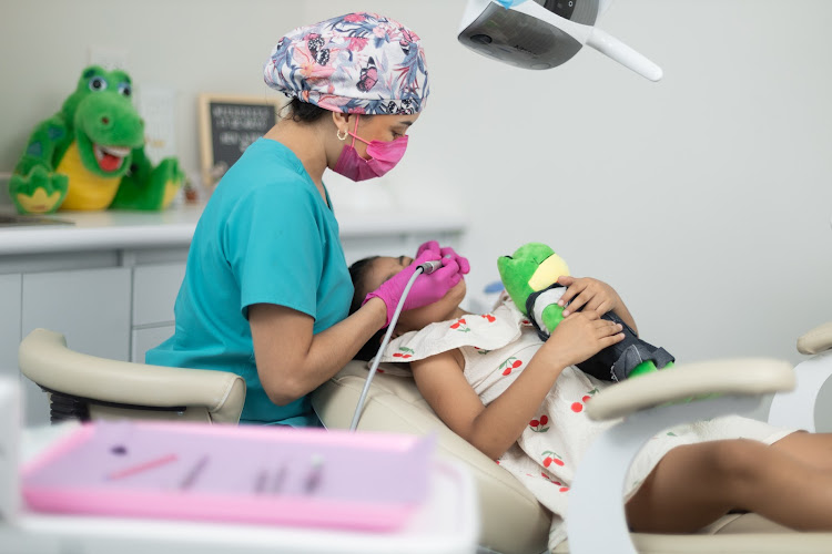 Dentista infantil en España - Un dentista con bata y mascarilla rosa atiende a un paciente infantil, que está tumbado en un sillón dental con un peluche verde en la mano. Al fondo se ven instrumentos dentales y un cocodrilo de juguete. | Clínica dental pediátrica Dentistainfantilcercademi